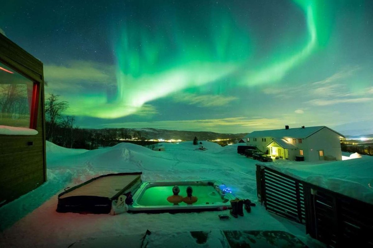 Two people relax in an outdoor hot tub near a hotel while watching the northern lights.