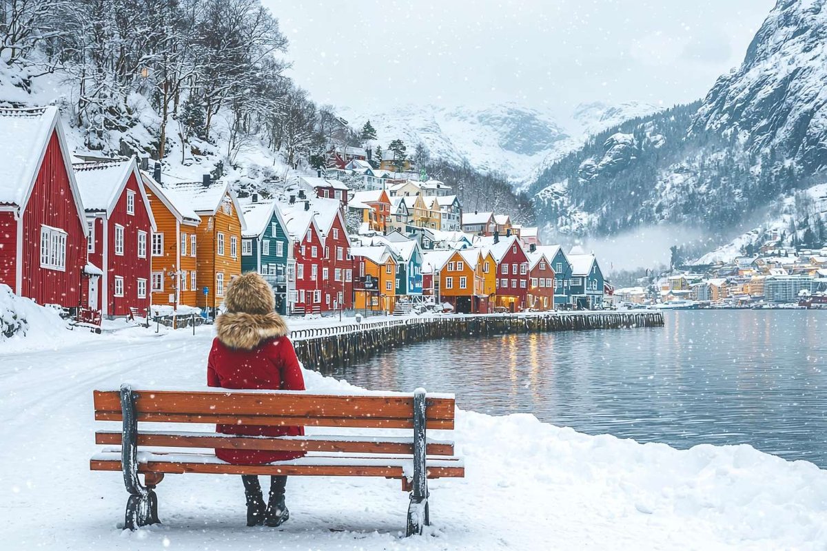 A woman sits on a bench by the water in a snowy Norwegian town.