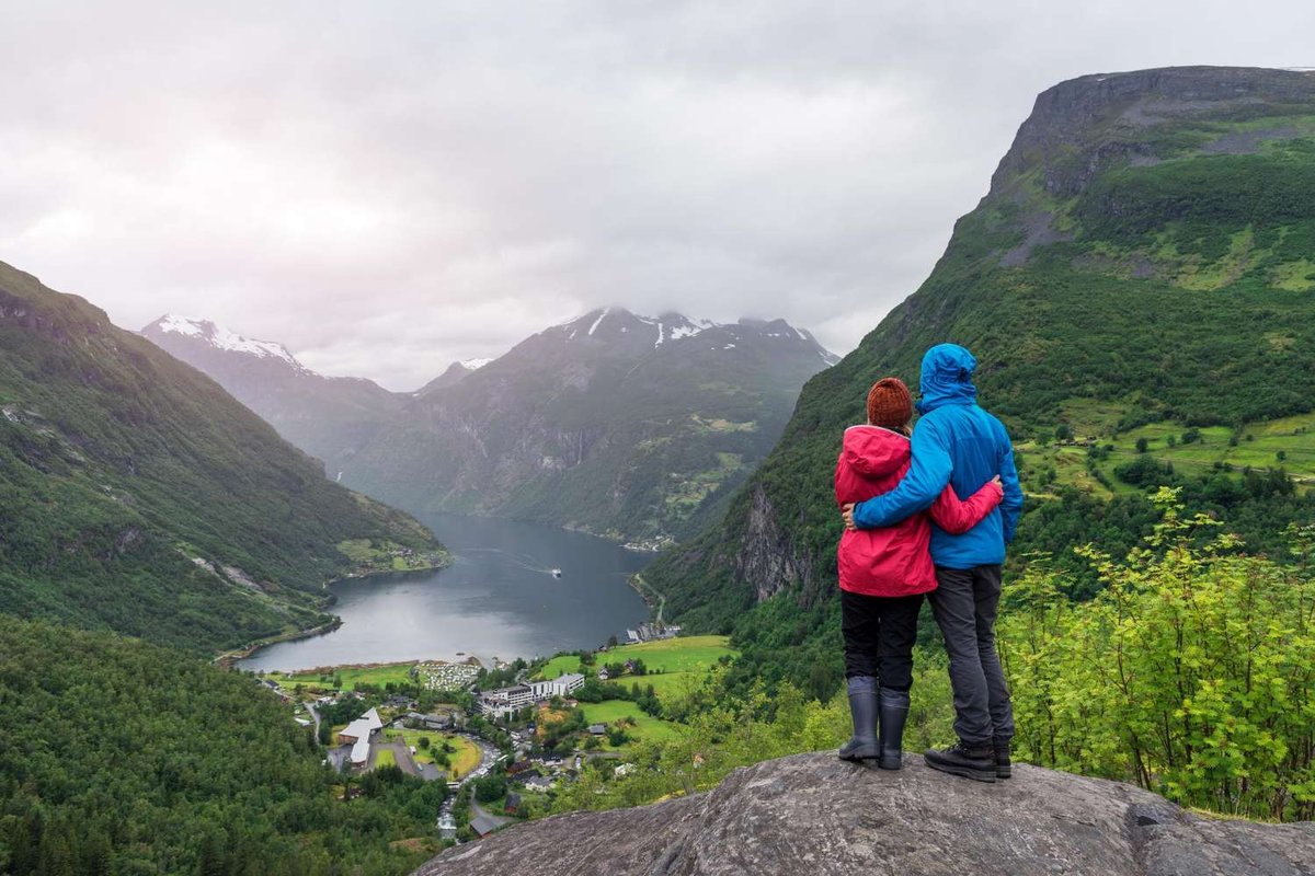 A couple in outdoor clothing looks at Geirangerfjord from a high viewpoint.