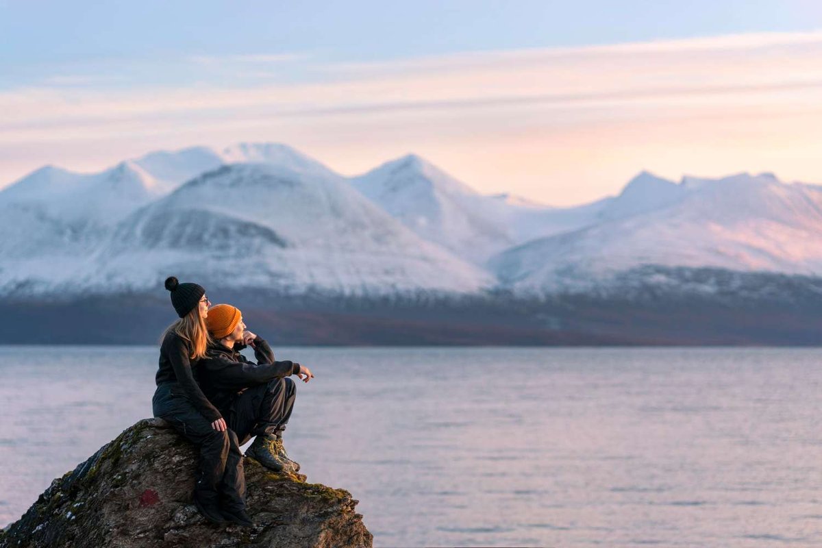 A couple sits on a rock by the sea overlooking snow-covered mountains.
