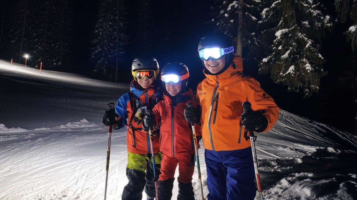 Three skiers in bright gear smile on Hafjell’s floodlit night slope—family vibe and photo-ready conditions.