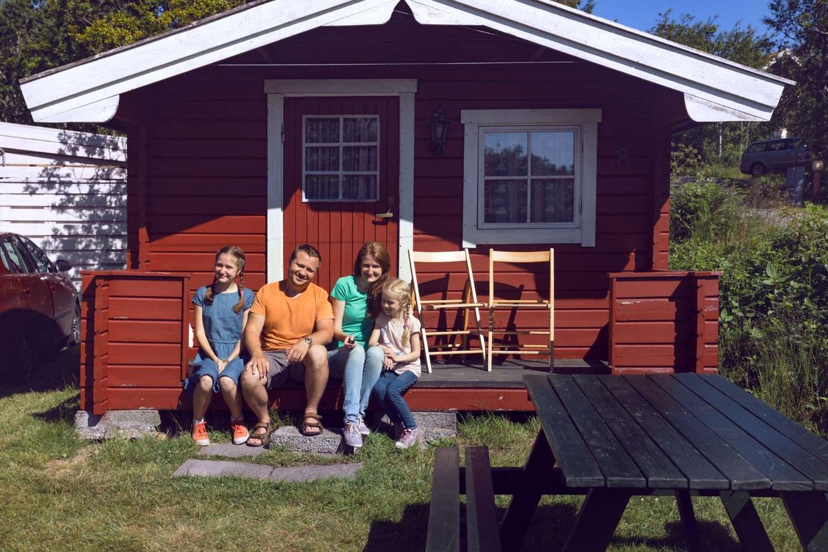 A family sits on the porch of a wooden guesthouse on a summer day.