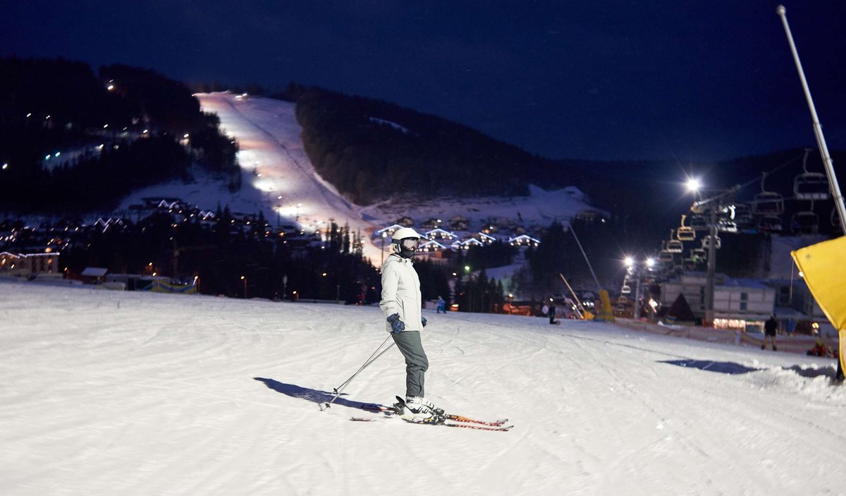 A skier on a floodlit evening slope with resort lights and chairlifts in the background.
