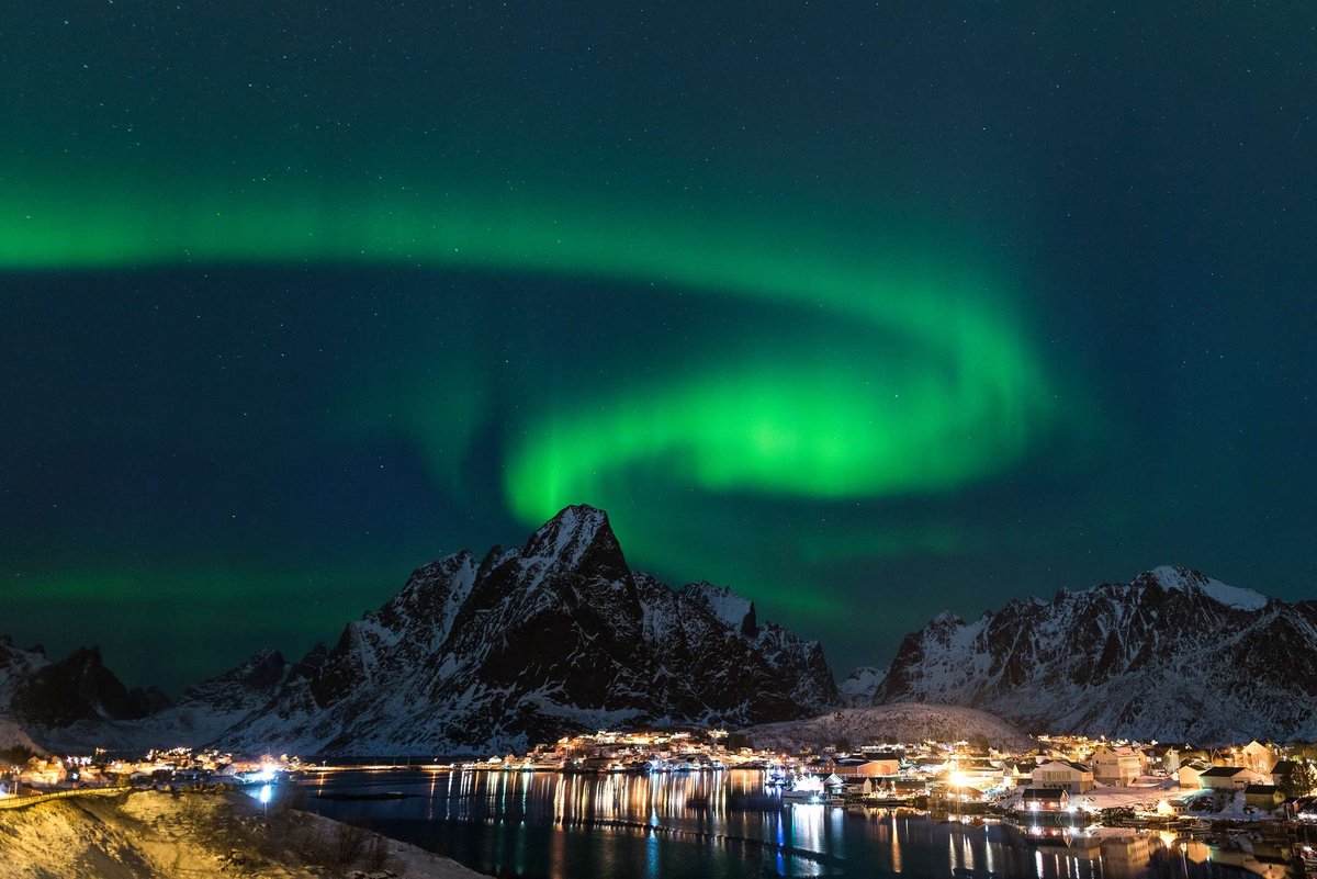 Bright Northern Lights illuminate the night sky over a snowy mountain and coastal village in Norway.