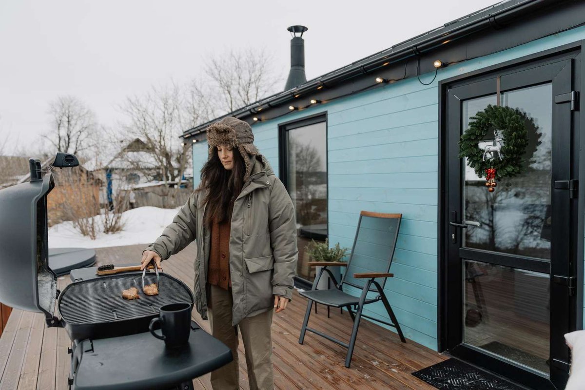A woman is grilling food next to a cabin at a winter campsite.