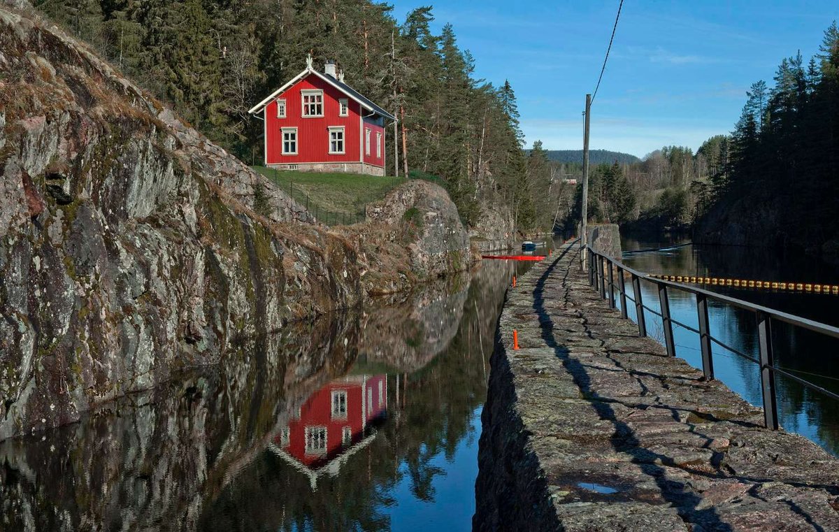 A red wooden house reflected in a still canal surrounded by rocky cliffs and pine trees in Norway.