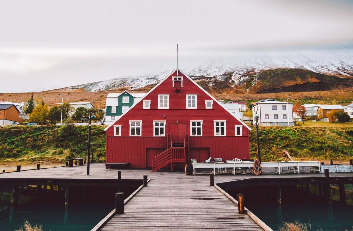A red wooden house with a dock is located by the shore, with hills and residential buildings in the background.